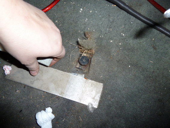 Hand reaching for a metal bracket on a dirty car floor near a seat bolt