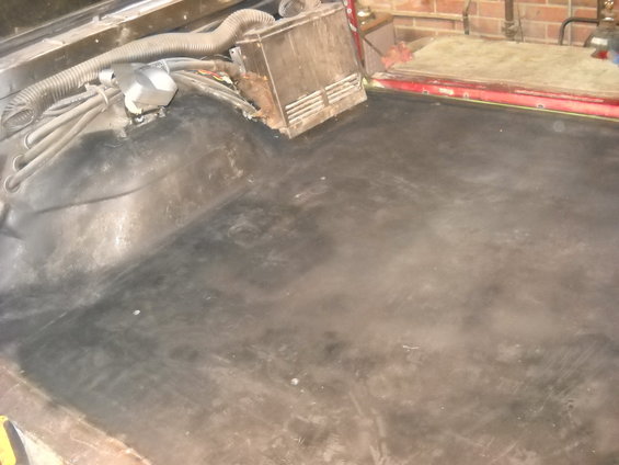 Vehicle interior floor with black carpeting and exposed wiring. A gray plastic housing with vents is visible