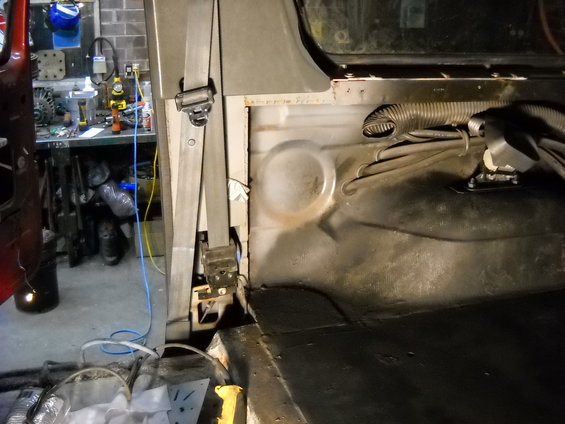 Vehicle interior showing a seatbelt mechanism and ribbed conduit. Tools and equipment are visible in the background