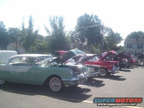 Several classic cars parked in a row, hoods open, at an outdoor event
