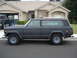 1977 Jeep Cherokee - A dark matte Jeep Wagoneer is parked on the street in front of a house, with another Jeep visible in the driveway.
