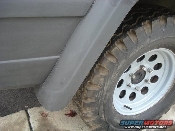 Close-up of a gray Jeep fender and off-road tire with a white rim. Supermotors.net logo visible