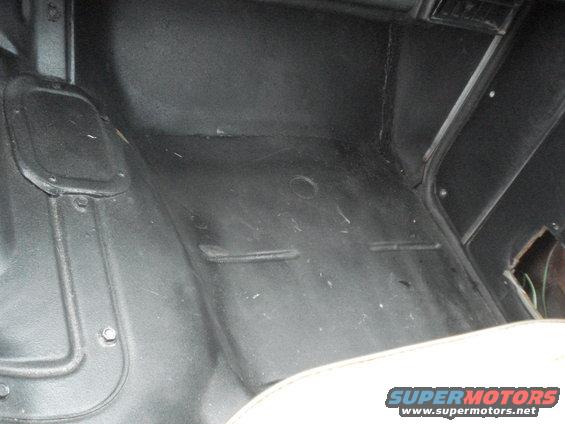 Black vehicle floor mat with raised ridges and a circular indentation. A white seat edge is visible at the bottom