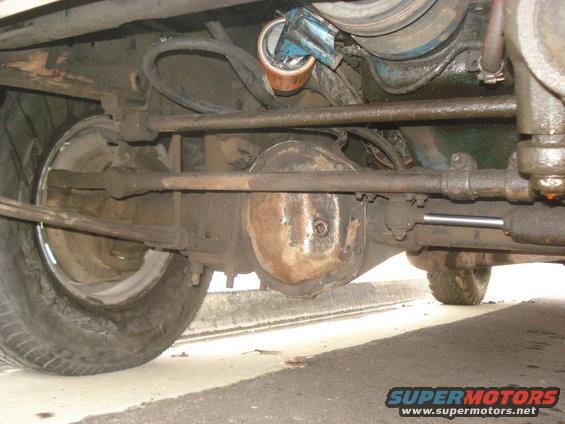 Underside view of a vehicle's rear axle, steering linkage, and tire. Shows rusty components and some fluid leaks