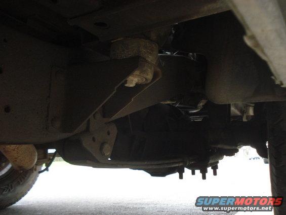 Underside view of vehicle frame, suspension, and drivetrain components. Tire visible on left
