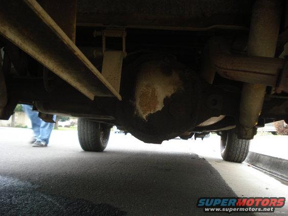 Underside view of a vehicle's rear axle and exhaust system. A person is visible in the background