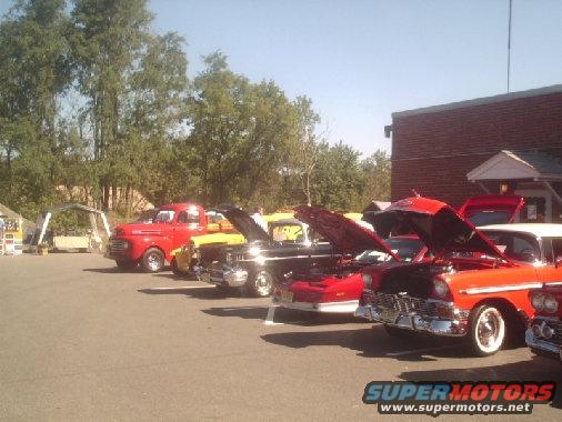 Red and white 1956 Chevrolet Bel Air with hood open, parked in a line with other classic cars