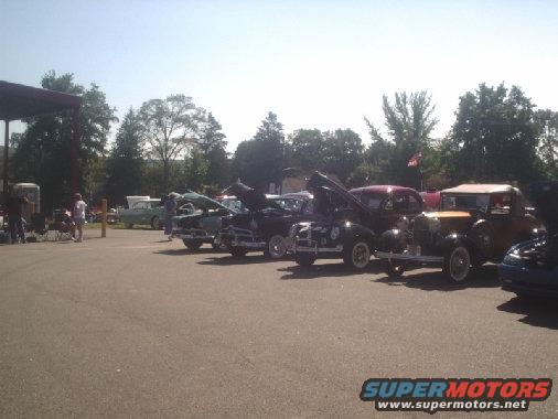 Several classic cars parked in a lot with hoods up, under a sunny sky