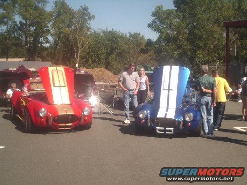 Two Cobra replicas, one red with gold stripes, one blue with white stripes, hoods open. People standing around them