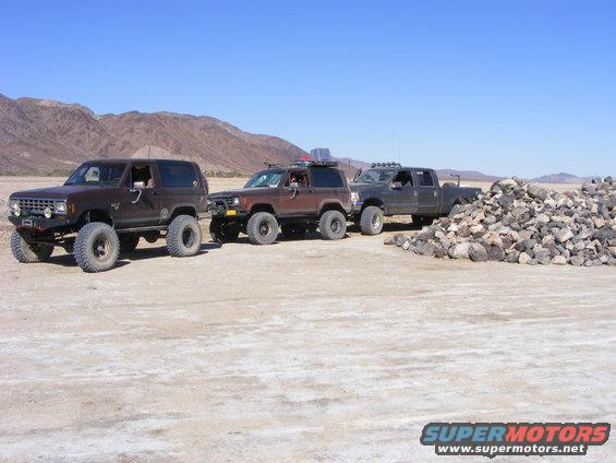 Three modified off-road vehicles parked in a desert landscape. The first two are SUVs, the third is a pickup truck