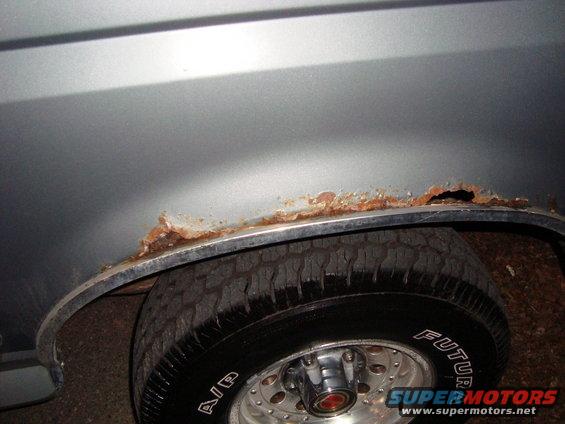 Close-up of a rusted-out rear wheel well on a vehicle. Tire is visible with 'FUTURE' written on it