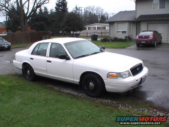White 2003 Ford Crown Victoria parked on a gravel and grass area in front of a house. A red sedan is parked in the driveway