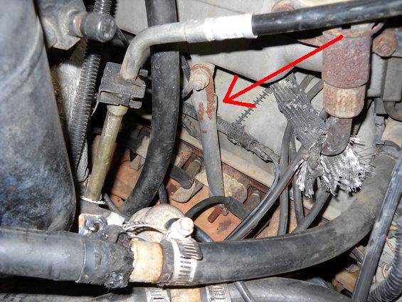 Close-up of engine bay showing hoses, metal lines, and wiring. A red arrow points to a rusty metal line