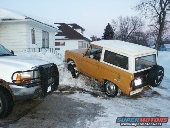 Yellow 1966 Ford Bronco with a white hardtop stuck in snow, viewed from the front 3/4 angle