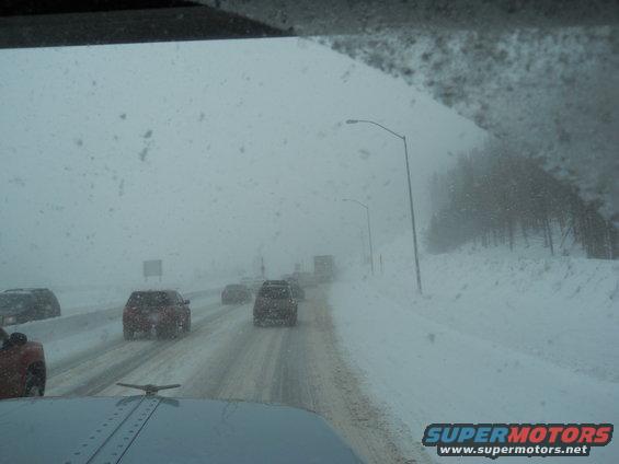 Truck cab interior view of highway traffic during a snowstorm. Several cars and trucks are visible on the snow-covered road