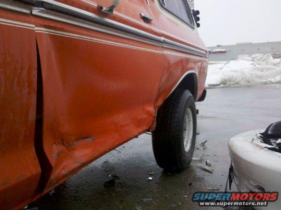 Orange SUV rear quarter panel with significant damage, next to a white vehicle. Broken glass on wet ground