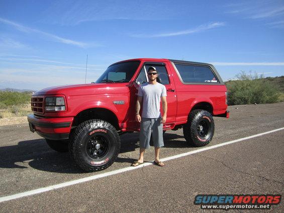 1996 Ford Bronco, red, with a man standing beside it on a paved road