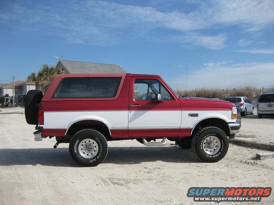 1995 Ford Bronco, two-tone red and white, side view, spare tire on rear door, parked on sand with buildings and sky in background