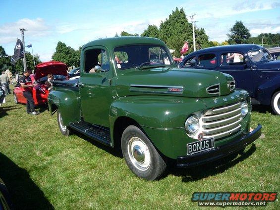 Green 1948 Ford F1 pickup truck with "BONUS 1" license plate. Another car is parked to the right