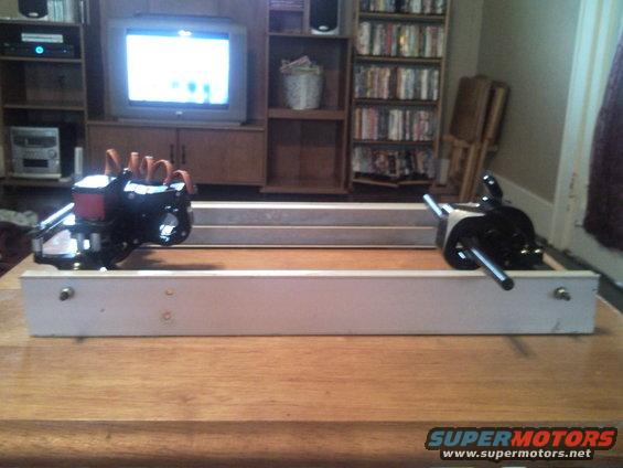 Engine components mounted on a wooden frame on a table. Supermotors.net logo visible