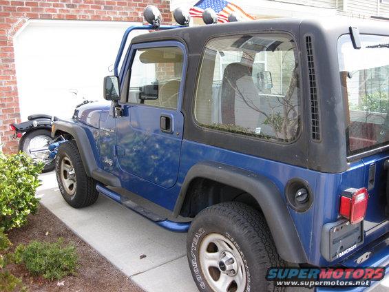 Blue Jeep Wrangler with black hardtop, roll cage, and auxiliary lights parked in driveway. Motorcycle visible behind it