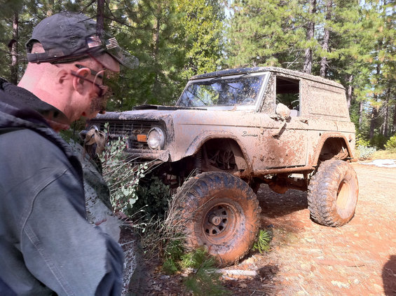 Muddy classic Ford Bronco with big tires, parked off-road with a person looking at it