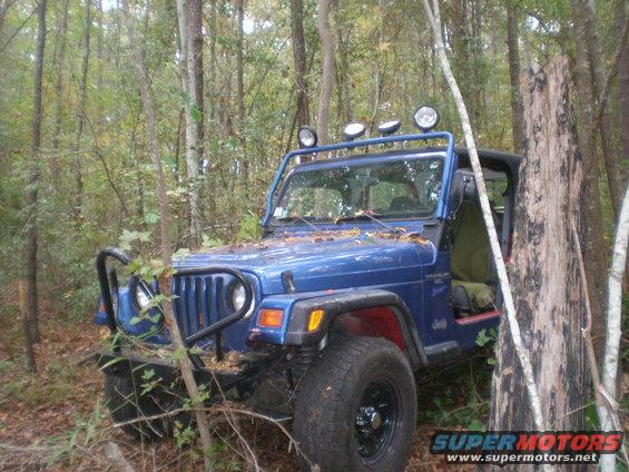 Blue Jeep Wrangler with aftermarket bumper and light bar parked off-road in a wooded area