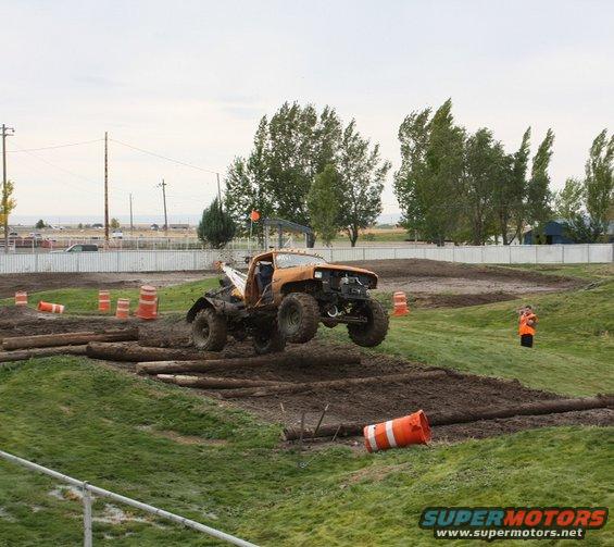 Orange off-road truck mid-air over logs on a dirt track