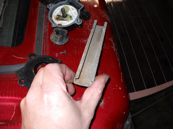 Hand holding a metal channel part next to disassembled water pump components on a red surface