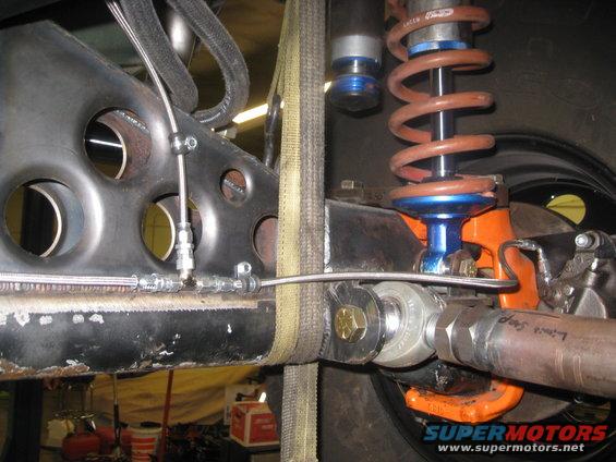 Vehicle suspension with orange shock absorber, brown coil spring, and black metal frame with circular cutouts. Straps secure the frame