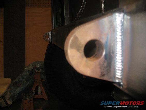 Close-up of a shiny metal bracket with a hole, welded to a larger metal piece, with a tire visible behind it