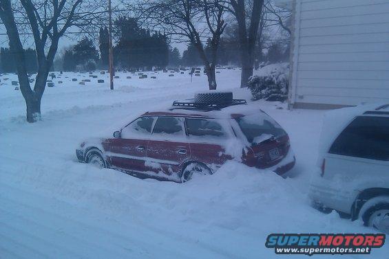 red station wagon with roof rack and spare tire stuck in deep snow next to a white minivan