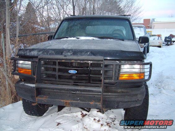 Front view of a black Ford Bronco with a brush guard, parked in the snow. Another vehicle is visible behind it