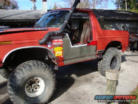 Red SUV with custom half doors, snorkel, and large off-road tires. Appears to be a Ford Bronco from the 1980s
