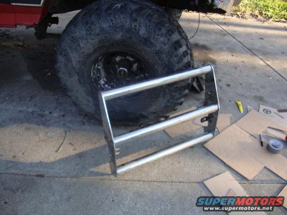 Chrome grille guard next to a large off-road tire on a red vehicle. Cardboard and tools are on the ground
