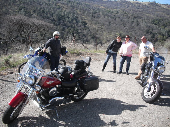 Three people stand near two motorcycles parked on a dirt road with hills in the background