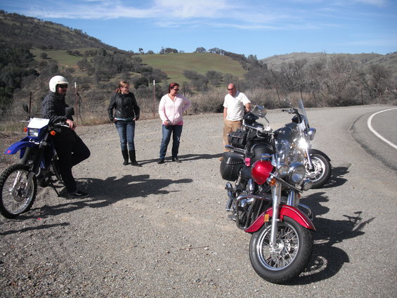 Two motorcycles parked on a gravel shoulder with four people standing nearby. Hills in background