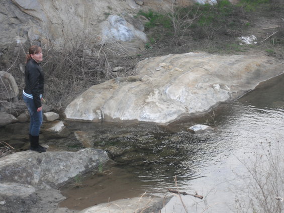Woman in black jacket and jeans stands on rocks next to a shallow stream with large boulders and dry vegetation