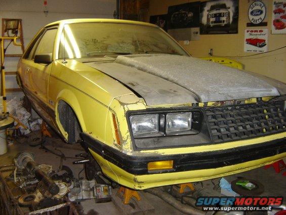 Yellow 1979 Ford Mustang coupe on jack stands in a garage, hood is gray primer