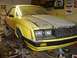 1979 Ford Mustang - A yellow 1979 Ford Mustang coupe is elevated on jack stands in a garage, showing its front end with a gray primer hood.