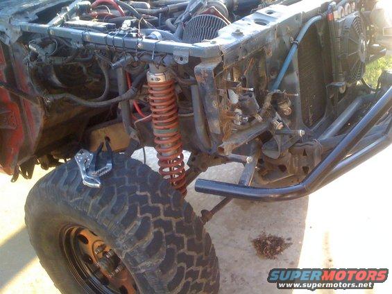 Disassembled vehicle front end with exposed engine components, red coilover shocks, and large off-road tire. A Vise-Grip is on the tire