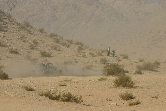 Off-road buggy kicking up dust while racing on a desert course. Two people in safety vests stand on a nearby hill