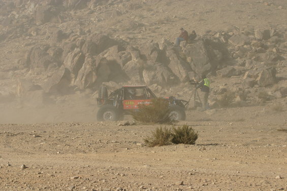 Off-road buggy kicking up dust on a desert course, with spectators on a rocky hillside