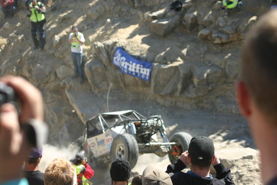 Off-road buggy with roll cage climbing a dusty, rocky incline. Spectators watch from the side. A banner reads 'Griffin Radiators'