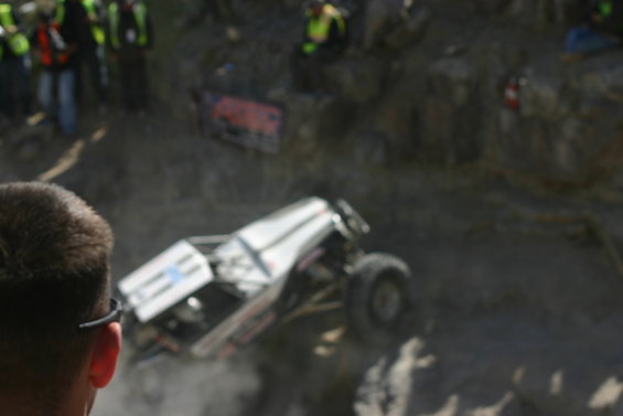 Off-road buggy with white body and roll cage climbing a steep, rocky incline. Spectators are visible on the left