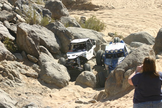 Two off-road buggies climbing over rocks in a desert landscape. A person watches from the side