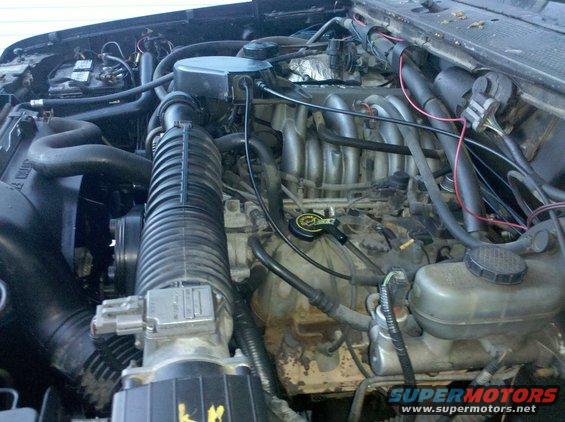 Engine bay with hoses, wires, and engine components visible. Some yellow markings on a black part