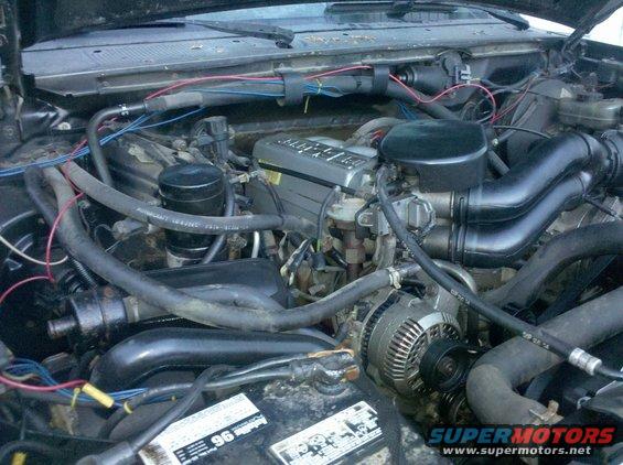 Engine bay with hoses, wires, alternator, and engine cover. Some text visible on battery