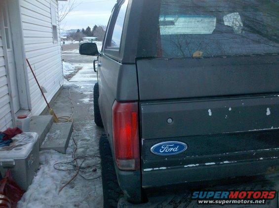Rear view of a dark-colored Ford Bronco parked next to a house. The Ford logo is visible on the tailgate