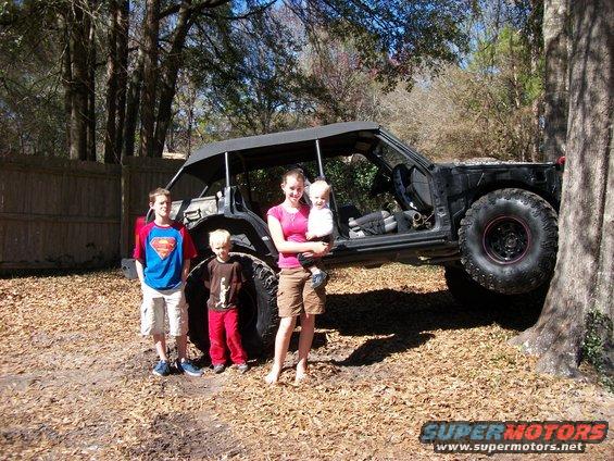 Modified off-road vehicle with a soft top and large tires, parked outdoors with children
